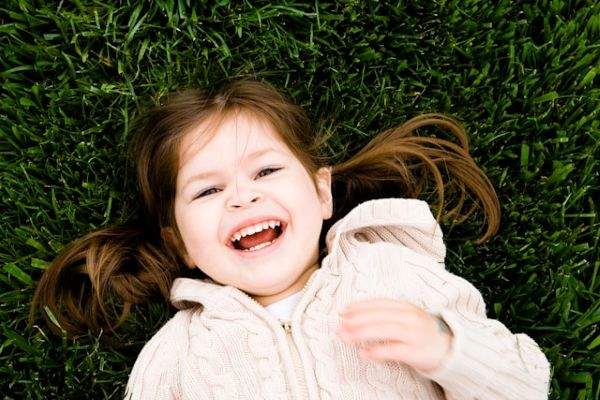 Niña de cabello castaño y pelo largo, tumbada sobre el césped verde, riendo felizmente