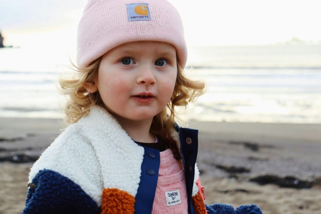 Niña rubia con gorro rosa y chaqueta mirando a cámara en la playa
