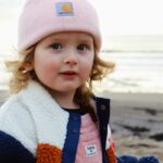 Niña pequeña con gorro rosa y chaqueta, mirando a cámara en una playa de clima frío