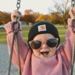 Niña feliz en un columpio con gafas de sol, gorro negro y jersey rosa de lana