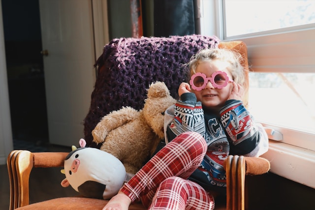 Niña con gafas rosas de flores sentada en una butaca con sus peluches