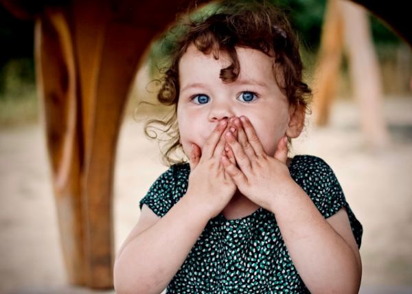 Niña pequeña con ojos azules y pelo rizado, vestida con vestido, mirando a la cámara con expresión de sorpresa y manos cubriendo la boca