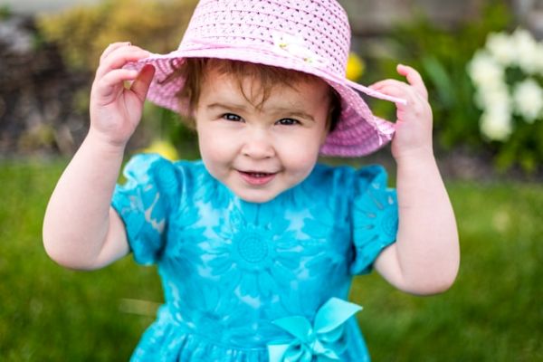Niña pequeña con vestido turquesa y sombrero rosa con flores, mirado a la cámara y sujetando el sombrero con las manos