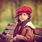 Niño con boina roja y bufanda sentado en un banco en un entorno otoñal
