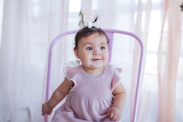 Bebé niña con vestido lila y lazo blanco en el pelo sentada en una silla lila